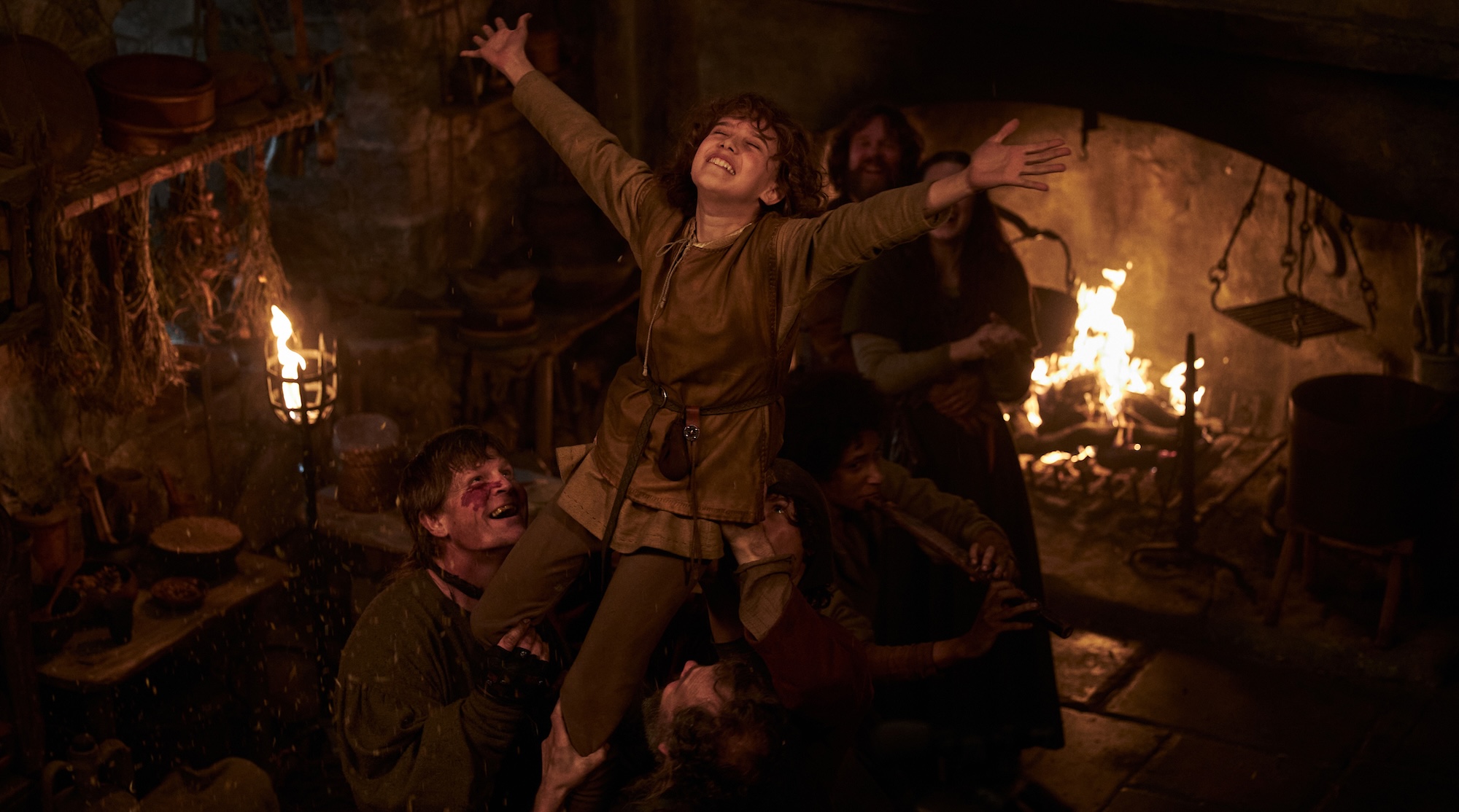 Abb. 2: Ronja (Kerstin Linden, Mitte) fühlt sich in der Mattisburg pudelwohl, in der es sie an nichts mangelt. Mit warmen dunklen Farben, die eine Gemütlichkeit ausstrahlen, wird das wilde und harmonische Leben der Mattisräuber auf der Burg dargestellt. Pressefoto: 2023 Viaplay Group/Filmlance International AB/Film i Väst/Ahil Films/ARD Degeto Film/Audrius Solominas. Die erste Staffel der Ronja Räubertochter-Serie ist in der ARD Mediathek bis zum 20.12.2025 verfügbar.